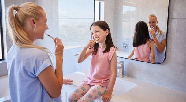 Adorable little girl getting ready with her mother as they brush their teeth with toothbrushes in the morning. Cute little daughter looking up to mother setting good example about oral hygiene and - Fotó, kép