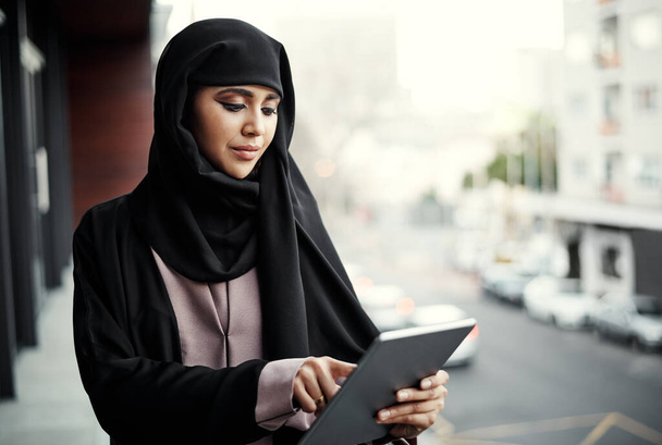 an attractive young businesswoman dressed in Islamic traditional clothing using a tablet while standing on her office balcony. - Foto, Imagen