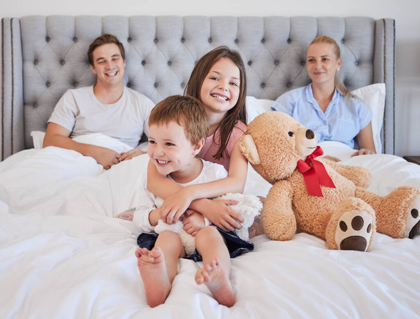 Adorable little girl embracing her younger brother with her teddybear next to her while sitting on a bed with per parents in the background. Loving young caucasian family bonding and spending time - Foto, Bild