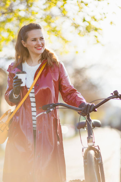 happy modern middle aged woman in red rain coat with bicycle and cup of tea walking outside in the city. - Foto, imagen