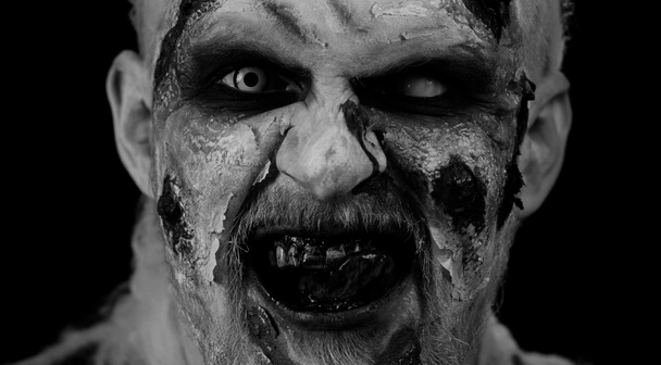 Nahaufnahme des Zombie-Mannes mit Make-up, Wundnarben und weißen Kontaktlinsen, der in die Kamera schaut und auf seine Zähne klickt, um im dunklen Raum Angst zu verbreiten. Finsterer Toter. Halloween, Dreharbeiten - Foto, Bild