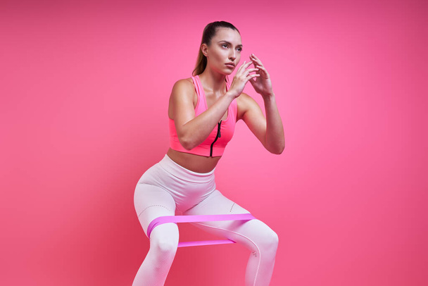 Confident young woman exercising with elastic band against pink background - Fotoğraf, Görsel
