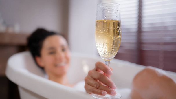 Out of focus young happy relaxed woman lying in foam bath holding glass champagne funny playful lady blowing on foam in bathroom enjoying flavorous alcoholic drink luxurious recreation for weekend - Photo, Image