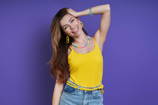 Attractive young woman holding hand in hair and smiling while standing against purple background - Foto, Imagen