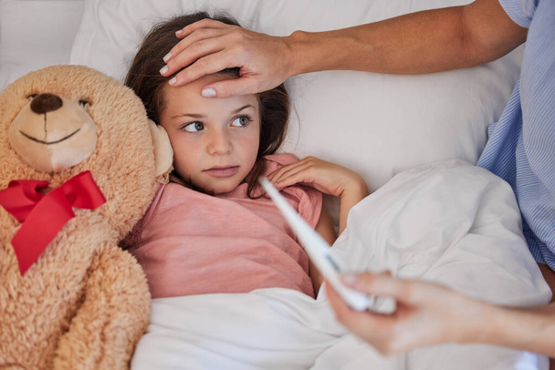 Sick little girl in bed with her teddybear while her mother uses a thermometer to check her temperature. Young concerned single parent sitting with sick child while and feeling her forehead . Small - Φωτογραφία, εικόνα