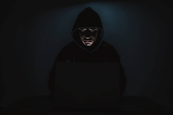 Egy hacker hosszú ujjú inget visel csuklyával a fején. Ül egy sötét szobában egy laptop segítségével A koncepció a személyazonosság lopás az internetes tábor hálózaton. fénymásolási hellyel. - Fotó, kép