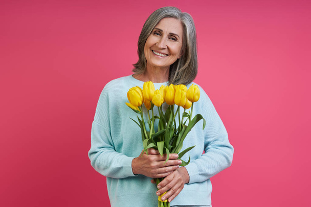 Happy senior woman holding bunch of tulips while standing against pink background - Фото, изображение