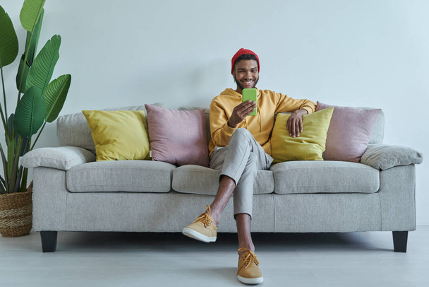 Happy African man looking at his smart phone while sitting on the couch - Foto, afbeelding