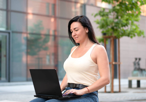 People. A beautiful young Caucasian girl in a light T-shirt is sitting with a black laptop in a park on a bench. Communication online. Rata online. Concept. Soft focus. - Foto, Bild