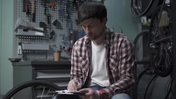 Male bicycle mechanic calculates cost of repairing a cycle with pen and clipboard in hand and yawns wearily and drinks coffee in bike shop. Using tablet while fixing bicycle. Tired repairman at work - Materiał filmowy, wideo