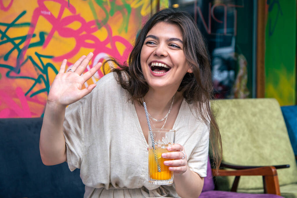 Ευτυχισμένη νεαρή γυναίκα με ένα ποτήρι λεμονάδα στο φόντο ενός φωτεινού ζωγραφισμένου τοίχου, το κορίτσι απολαμβάνει ένα καλοκαιρινό ποτό. - Φωτογραφία, εικόνα