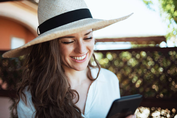smiling trendy woman in white shirt with hat using smartphone applications in the patio of guest house hotel. - Photo, image