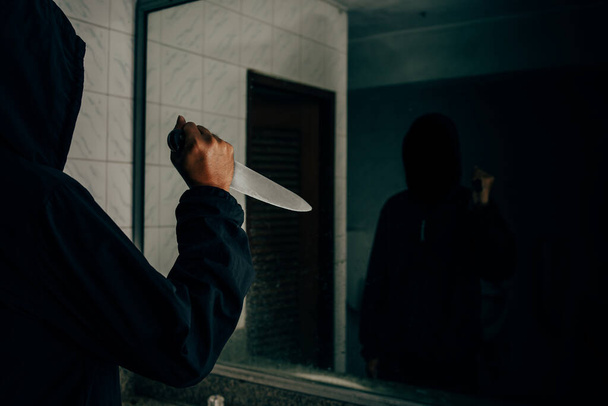 Vista trasera de un asesino sosteniendo un cuchillo En el baño, se miraba a sí mismo en el espejo. - Foto, Imagen
