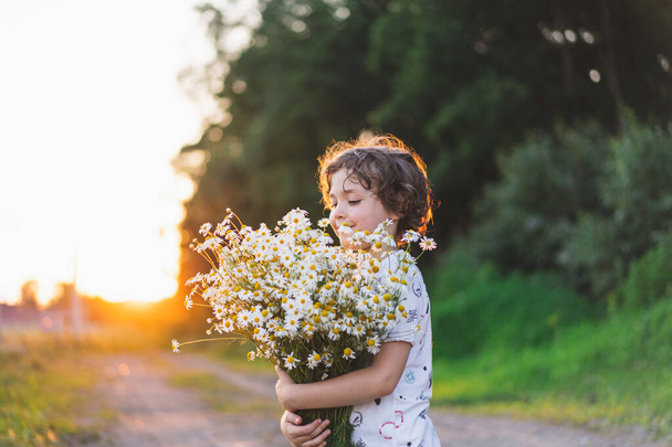 Cute smiling boy at camomile field at sunset in soft sunlight. Life without allergies, breathe freely. Boy and daisies. Child dreaming and smiling against the background of a camomile field. - Fotó, kép