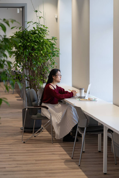 Pensive aasialainen nuori nainen etsii harkitusti kannettavan tietokoneen näytön työskennellessään etänä nykyaikaisessa julkisessa kirjastossa, japanilainen naispuolinen opiskelija käyttää internetiä opetustarkoituksiin. Etäopetuksen käsite - Valokuva, kuva