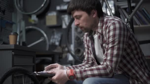 Tired bicycle mechanic drinks coffee at his workplace while calculating cost of working in bike shop. Overtime work. Boring paperwork. Inventorying items. Man calculates price of repairing the bike. - Materiał filmowy, wideo