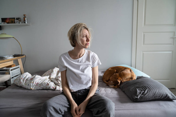 Mental disorders in mid-life. Unhappy depressed middle-aged woman living alone with dog sit on bed looking into distance, thinking about personal problems, feeling lonely, suffering from depression - Foto, afbeelding