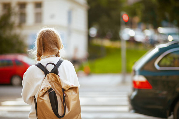 Seen from behind trendy school girl in white sweatshirt with backpack crossing crosswalk and going to school outdoors in the city. - 写真・画像