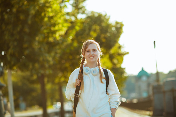 smiling stylish girl in white sweatshirt with backpack and headphones going to school outdoors in the city. - Foto, Imagem