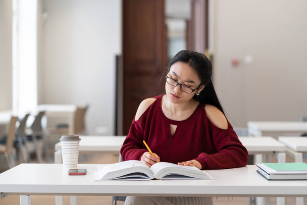 Young asian girl student making assessments from book for study. Female preparing for exam in university, college or high school, make homework sitting in empty classroom or library. Education concept - Photo, image