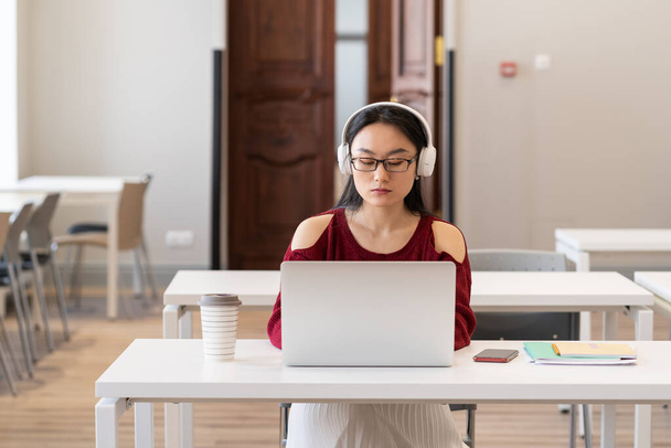 Vakava aasialainen college tyttö kuulokkeet katsella video oppitunti tai webinar Internetin kautta kannettavan tietokoneen istuessaan tyhjässä lukusalissa, naispuolinen opiskelija työskentelee valmistumisen projekti kirjastossa - Valokuva, kuva