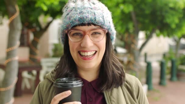 Happy woman enjoying coffee while out traveling in the urban city. Portrait of an edgy young woman wearing glasses and a beanie and having a positive attitude while exploring and sightseeing downtown. - Video