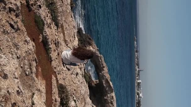 A young carefree woman from behind is sitting alone at the edge near sea caves love bridge in Ayia Napa, Cyprus - Footage, Video
