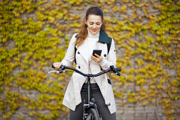 smiling modern woman in beige trench coat with bicycle sending text message using smartphone against the green wall outdoors on the city street. - Foto, afbeelding