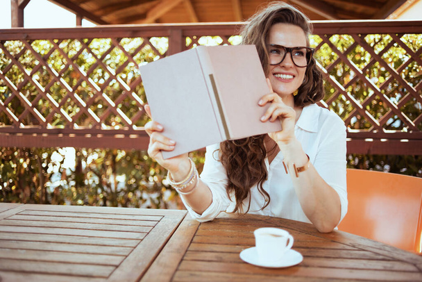 happy stylish middle aged housewife in white shirt with cup of coffee, book and eyeglasses sitting at the table in the patio. - Photo, image