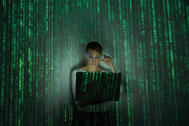 Confident woman adjusting futuristic glasses while using laptop against dark background - Foto, afbeelding