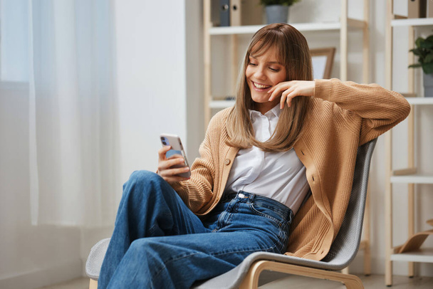 Cute młoda blondynka w ciepłym swetrze robi selfie używać telefonu trzymać za rękę w pobliżu twarzy siedzi w fotelu w nowoczesnym domu. Przerwa w pracy, przerwa, media społecznościowe w koncepcji wolnego czasu. Przestrzeń kopiowania - Zdjęcie, obraz