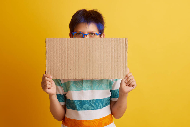 ένα αγόρι με γυαλιά σε κίτρινο φόντο με διαφορετικά συναισθήματα, κρατά ένα χαρτονένιο πανό με κενό χώρο για κείμενο - Φωτογραφία, εικόνα