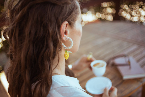 Смотреть из-за современной женщины в белой рубашке с чашкой кофе, книги и очков, сидя за столом в патио гостевого дома отеля. - Фото, изображение