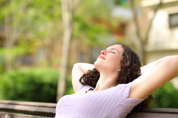 Relaxed woman resting with arms on head sitting in a bench in a park - Foto, Imagen