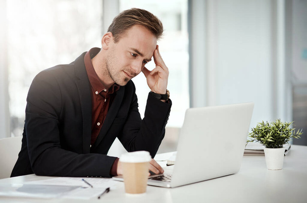 egy fiatal üzletember, aki stresszesnek tűnik, miközben egy laptopon dolgozik egy irodában.. - Fotó, kép