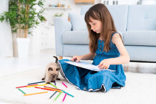 Child girl painting with kitten and sitting on the floor. Little female person drawing and playful kitten next to her at home playing with colorful pencils. High quality photo - Foto, afbeelding
