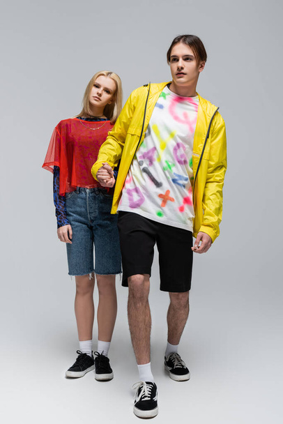 Full length of stylish man in windbreaker holding hand of girlfriend on grey background - Foto, immagini