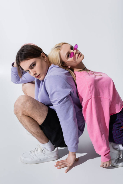 Stylish young couple looking at camera while sitting back to back on grey background - Zdjęcie, obraz
