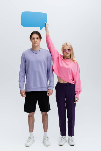 Full length of woman in sunglasses holding speech bubble above boyfriend on grey background - Foto, immagini