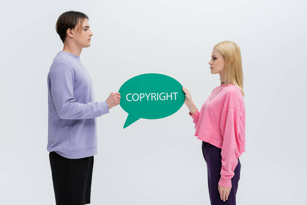 Vue latérale du jeune couple en sweat-shirts tenant une bulle d'expression avec lettrage copyright isolé sur gris  - Photo, image