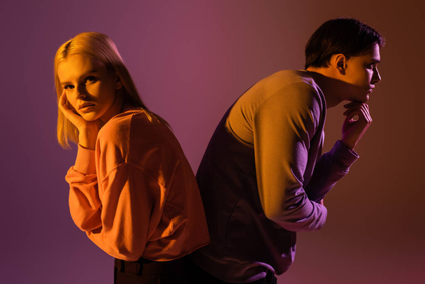 Young stylish couple in sweatshirts posing back to back on purple background with lighting  - Fotografie, Obrázek
