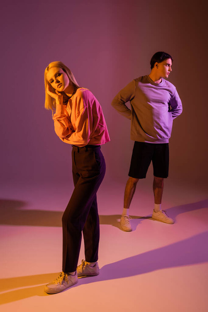 Young blonde woman looking at camera near brunette boyfriend on purple background with lighting  - Foto, afbeelding