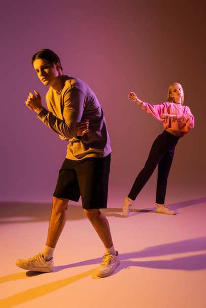 Full length of stylish couple in sweatshirts and sneakers on purple background with lighting  - Фото, изображение