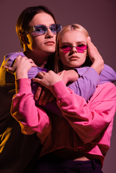 Stylowa para w okularach przeciwsłonecznych przytula się i patrzy na kamerę odizolowaną na fioletowo z oświetleniem  - Zdjęcie, obraz