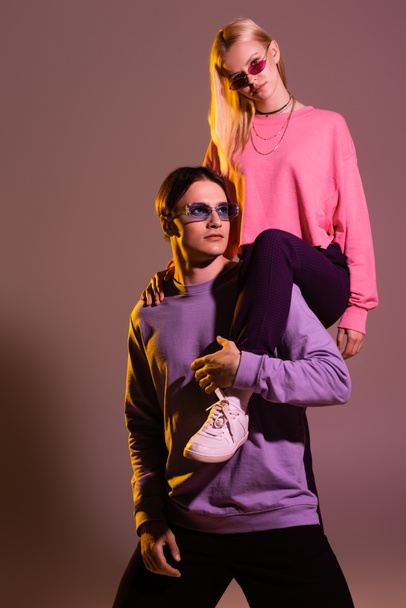 Stylish woman in sunglasses posing with boyfriend on purple background with lighting  - 写真・画像