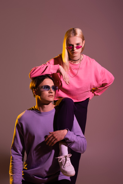 Pretty blonde woman looking at camera near boyfriend in sunglasses on purple background with lighting  - Zdjęcie, obraz