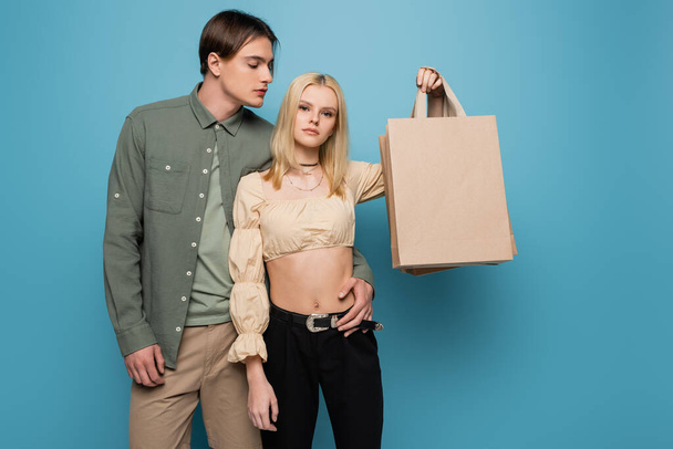 Stylish man hugging girlfriend with shopping bags on blue background  - Fotografie, Obrázek