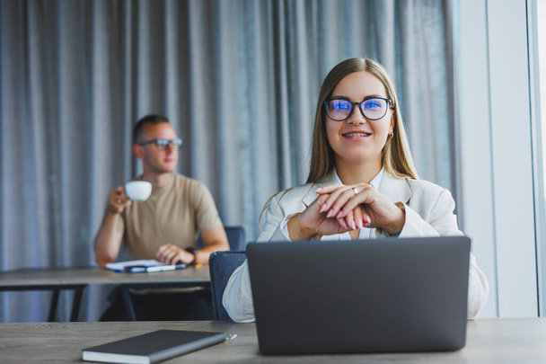 Attraktive junge Frau mit Brille sitzt an einem Tisch mit Laptop in einem Coworking Space und diskutiert mit einer Kollegin einen Projektplan. Arbeitsablauf im Büro. - Foto, Bild
