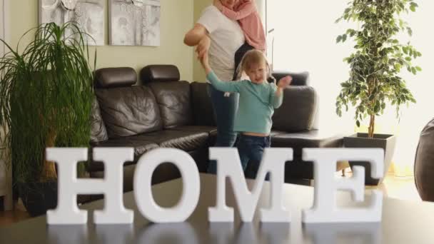 Aktivní maminka hlídání dětí a roztomilé dcerky skákání v moderním obývacím pokoji, točení kolem v tanci na pozadí nápis HOME. Šťastná rodina - Záběry, video
