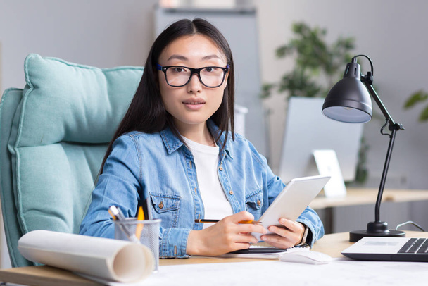 Retrato de una joven hermosa arquitecta y diseñadora asiática en gafas trabaja en un proyecto, se sienta en un escritorio, sostiene una tableta en sus manos. Mira a la cámara, sonríe. - Foto, imagen